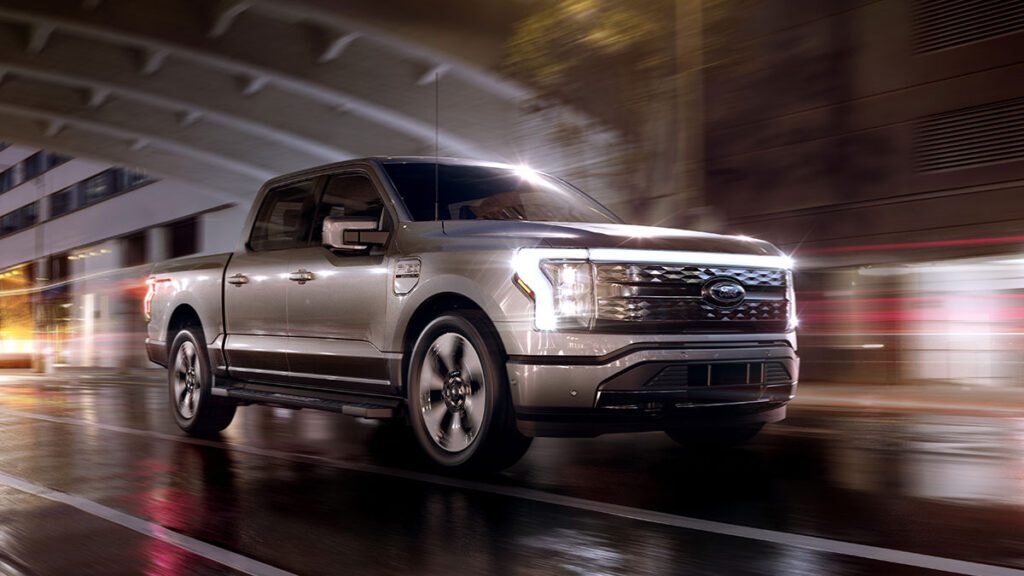 A silver pickup truck driving at night on a wet city street, under a bridge with blurred city lights.