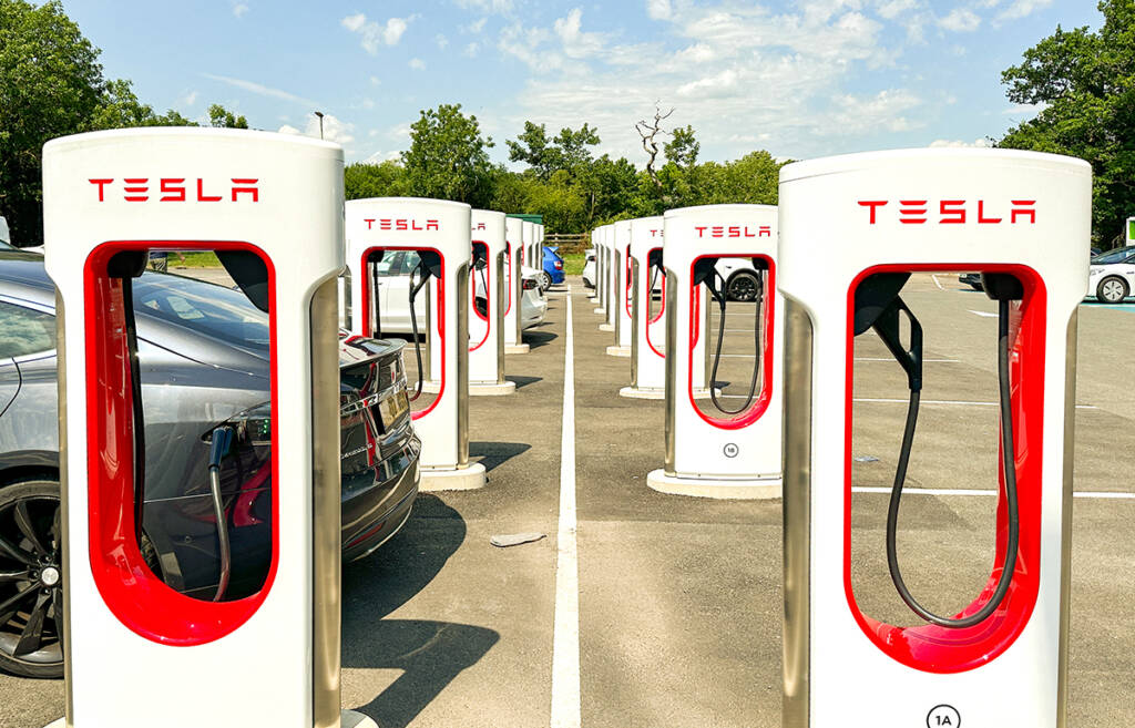 Tesla charging station with multiple chargers and parked cars in an outdoor parking lot beneath a clear sky.
