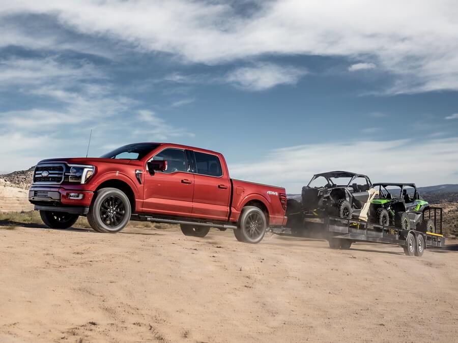 Red pickup truck towing two off-road vehicles on a trailer across a sandy landscape under a cloudy sky.