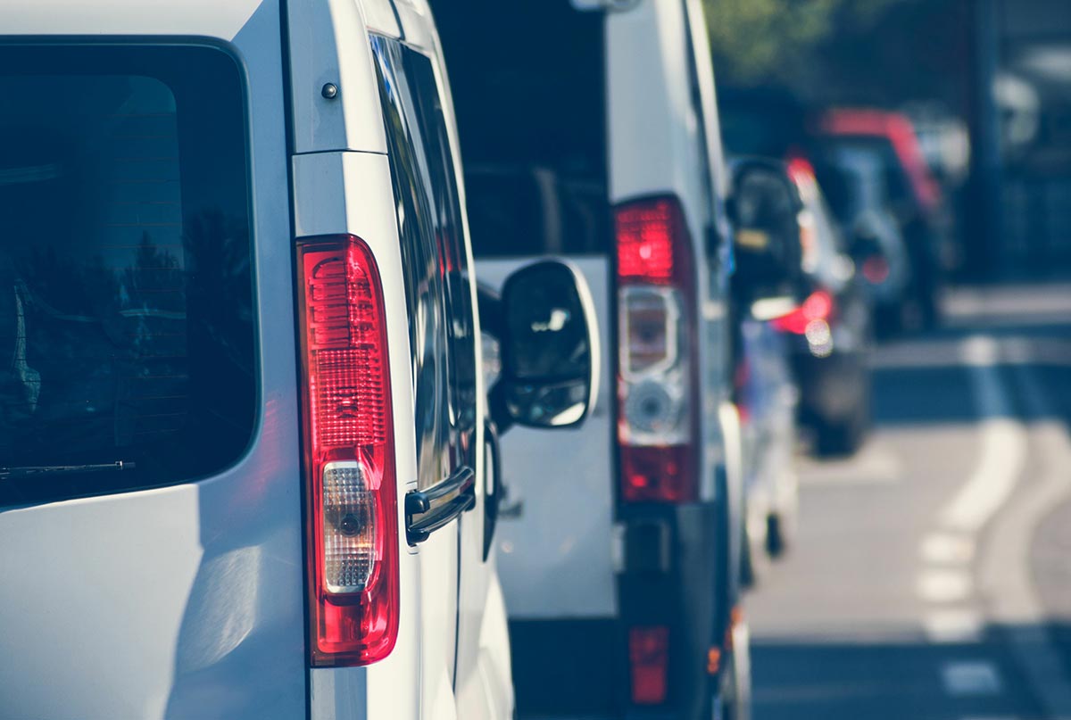 A line of white vans and cars in close proximity on a sunny road.