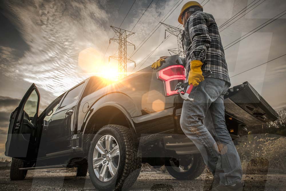 Worker in hard hat and gloves stands by pickup truck near power lines during sunset.